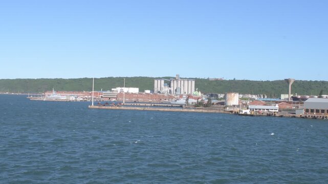 scenic wide water view across durban harbor towards maydon wharf industrial terminals, grain silos, and coastal port infrastructure backed by green coastal bluffs under a clear blue sky