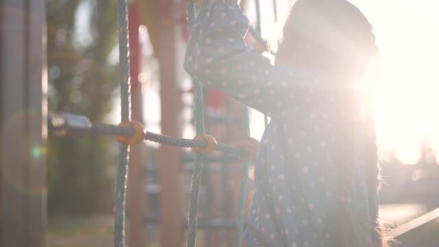 Girl climbing rope net on playground at sunset. Child grips rope ladder on climbing frame. Kid plays actively in park. Girl with braid climbs outdoor playground equipment. Active child enjoys summer.