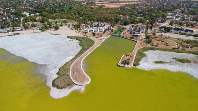 Drone view of Lake Velence in Velence, Hungary, showing shallow green water, exposed lakebed, reed-lined shore and a waterfront promenade during summer drought.