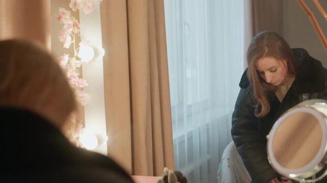 Woman adjusts her jacket in front of mirror with soft lighting and floral decor. Makeup brushes are visible on table suggesting preparation for event or outing