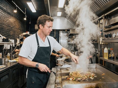 Chef cooking noodles and vegetables on a griddle in a busy kitchen with steam rising