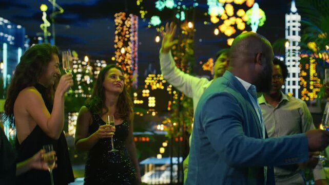 Close up of a happy man dancing with a drink at a New Year's Eve party, moving energetically to music with friends in a luxury rooftop setting as fireworks glow in the background at night.