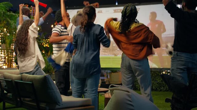 Close up of ecstatic football spectators hugging while watching a live match on a rooftop terrace screen, celebrating success and sharing joy during late-night sports viewing above the city.