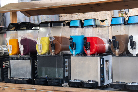 Multiple slush machines with various colorful frozen drinks are displayed on a wooden counter in an outdoor setting, ready for serving.