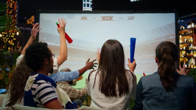 Friends watch a projected football match and react unhappily after conceding a goal, while frustrated fans respond to their team's loss during a live rooftop broadcast at night above the city skyline.