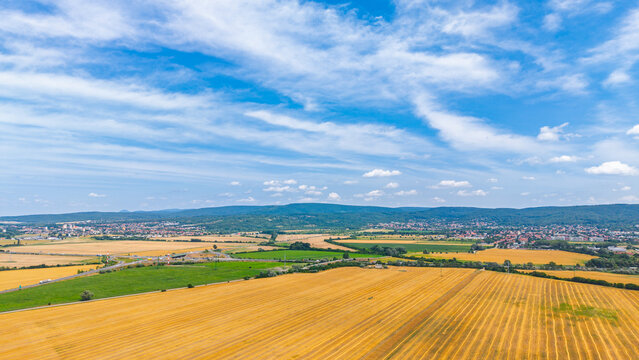 Aerial view of agricultural fields and distant hills under blue sky. Golden harvested fields spread across a rural landscape towards distant hills and towns under scattered clouds.