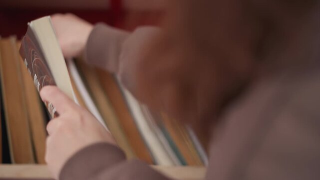 Person browses through various book titles picking up different volumes to examine. focus remains closely framed around interaction with printed materials against red background shelf