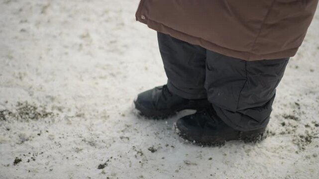 Close up shots show person legs and black rubber soled shoes with laces moving through light snow. ground is white slightly dirty patches visible. Footwear details like tread pattern are