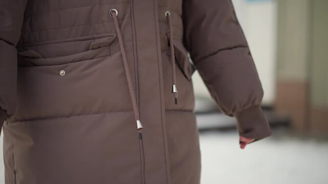 Brown winter coat focusing on details like pockets and drawstrings. fabric texture stitching seams zipper closure button fastenings are visible. Background suggests outdoor urban setting