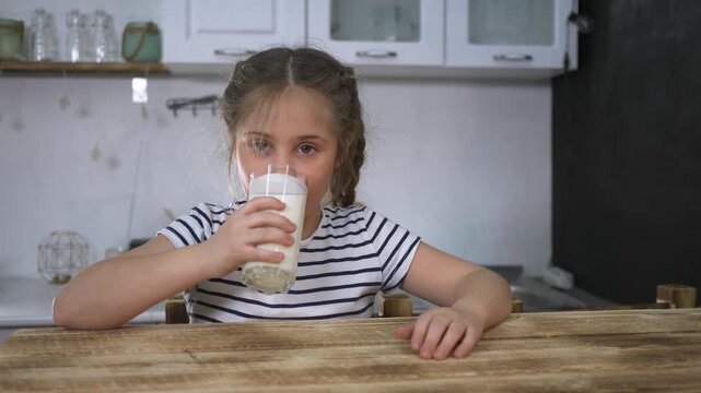 Girl drinking milk from glass in kitchen. Child sips milk at wooden table and smiles. Kid enjoys fresh milk beverage indoors. Happy girl holds glass of milk. Healthy drink for child at home kitchen.