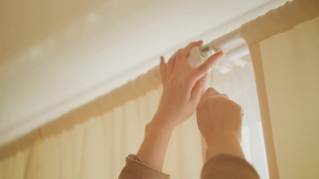 Woman hands adjust cream colored window blinds indoors under diffused lighting. close up view focuses on smooth texture of fabric and metal hardware as natural light filters