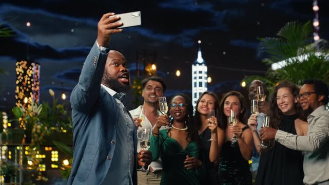 Smiling partygoers pose for smartphone selfies with champagne flutes during a rooftop celebration at night, raising sparkling wine glasses together while capturing a cheerful group photo.
