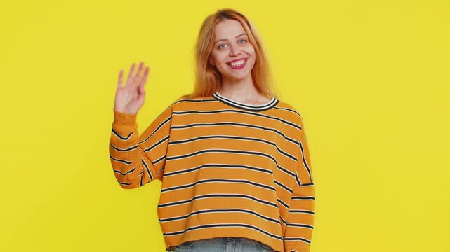 Young Caucasian woman smiling friendly at camera, waving hands gesturing hello hi greeting or goodbye welcoming with invitation hospitable expression. Happy girl isolated on yellow studio background