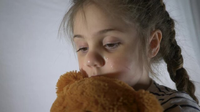 Sad child crying while hugging teddy bear. Little girl with tears on face holds stuffed toy. Crying kid wipes eyes with hand. Sad girl with braid embraces plush bear. Unhappy child shows emotion.