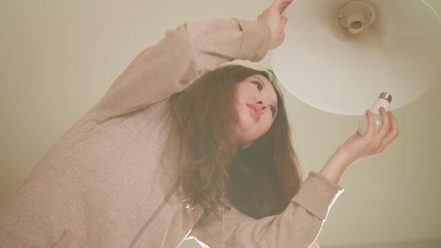 Young lady with long hair and casual clothing reaches up to change white LED light bulb inside ceiling lamp. Woman holds both hands around base of old or new bulbs tilts her head back