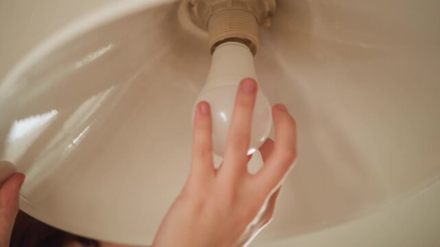 Close up view of woman hands adjusting light bulb in ceiling lamp. Hands carefully remove or position white glass shades around fixture focusing on precise movements