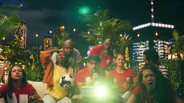 Unhappy football supporters react to a conceded goal while gathered on a rooftop terrace, showing frustration after a missed chance during a tense live soccer broadcast at night with friends nearby.