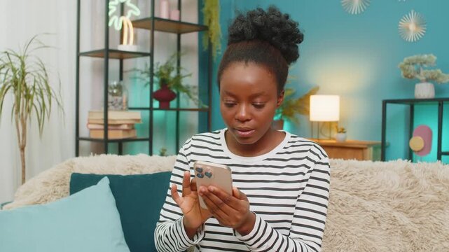 African American woman updating social media profile. Sitting on sofa at home, scrolling photos, writing descriptions, laughing at memes. Relaxed mood, casual interaction indoors. Enjoying time online