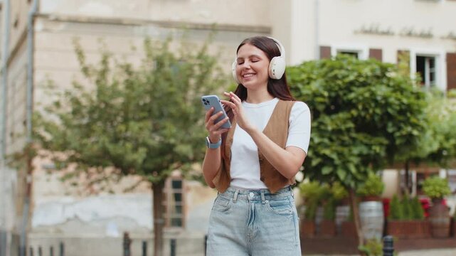Happy relaxed overjoyed young adult girl in wireless headphones choosing, listening favorite energetic disco rock n roll music dancing outdoors. Caucasian female woman on urban city street background.
