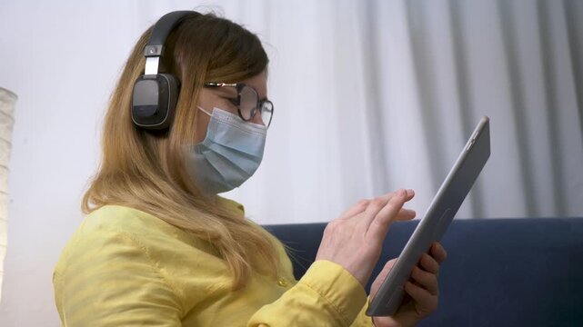 Woman in face mask uses tablet on sofa. Girl wearing headphone browses digital device at home. Masked woman with glasses scrolls tablet indoors. Quarantine lifestyle with mask and headphone on couch.