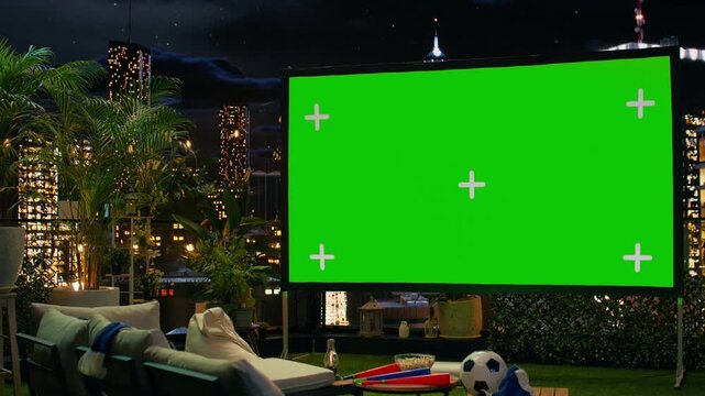 Green screen display is prepared for football match projection on a rooftop terrace overlooking the illuminated city skyline during nighttime sports viewing.