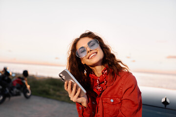 Obraz premium Young woman in a red jacket smiles while looking at a smartphone outdoors at sunset, with a calm water backdrop. Candid lifestyle shot for social, travel, or fashion use. Great for branding.