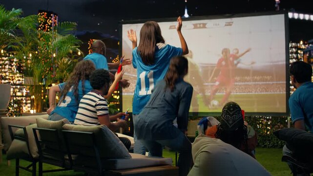 Soccer supporters watch a projected match on a rooftop and discuss a controversial referee call, feeling worried during a tense nighttime sports event.