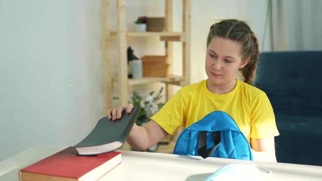 Girl packing school backpack with books at home. Child with Down syndrome puts notebook into bag at desk. Student prepares school supplies. Kid packs backpack for education. Girl organizes book stack.