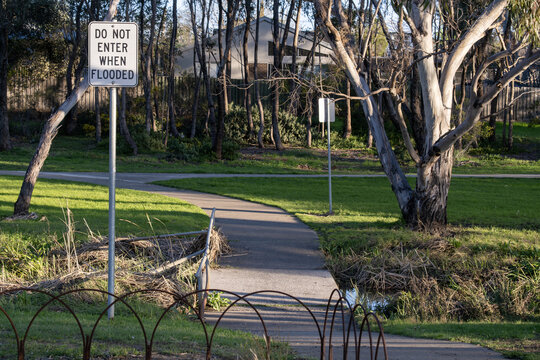 A flood warning sign stands beside a footpath that crosses low-lying ground near Laverton Creek, framed by tall native eucalyptus trees and parkland, residential houses nearby. Melbourne Australia