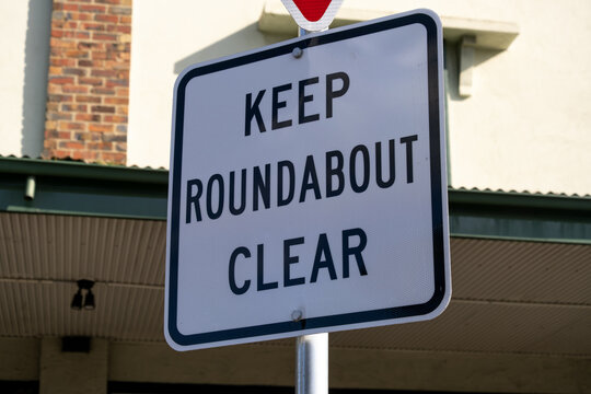 A white rectangular "Keep Roundabout Clear" traffic sign stands on a metal post in front of a commercial building in Melbourne Australia. Intersection management, Australian road rules