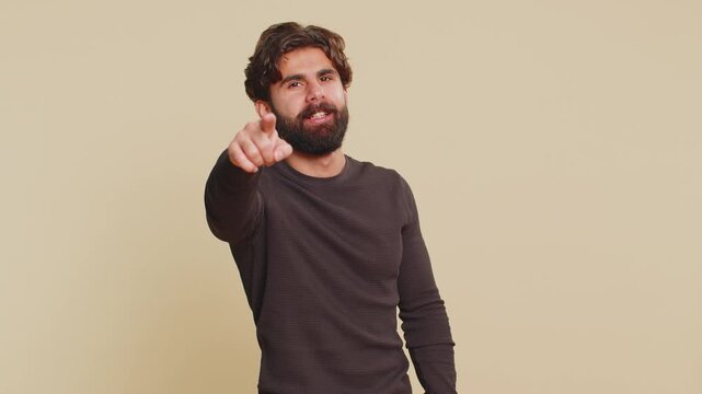 I choose you. Indian young man pointing to camera and looking with playful happy expression, making choice, showing direction. Hispanic Hindu guy smiling isolated alone on beige studio background.