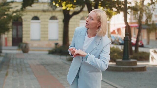 Serious sad businesswoman standing outdoors looking around waiting for business meeting impatient woman dissatisfied looks at wristwatch, checks time on watch annoyed by being late lateness concept.