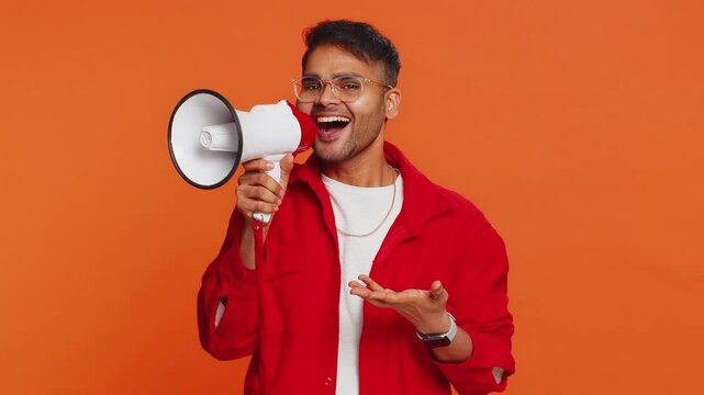 Indian young man talking with megaphone, proclaiming news, loudly announcing advertisement discounts sale, using loudspeaker to shout speech. Happy Arabian guy isolated on studio orange background