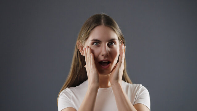 Young woman holding hands to her face expressing surprise and shock on a gray background