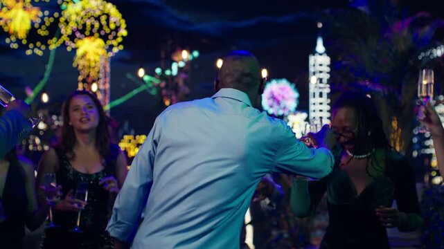 Close up of happy friends dancing at a late-night New Year's Eve party while partygoers hold champagne glasses and enjoy music at midnight.
