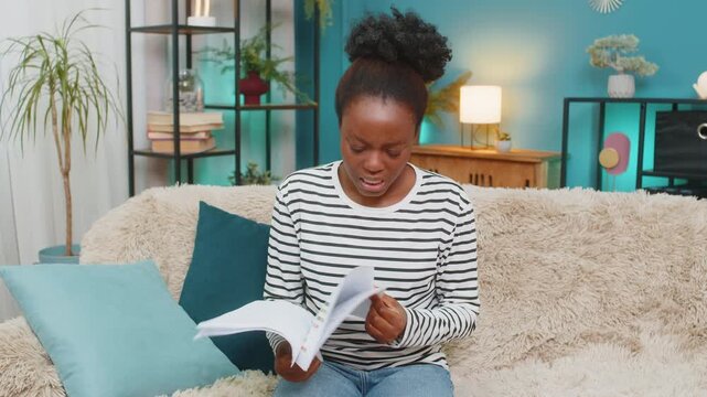 African American woman sitting on home sofa, feeling upset stressed as reviews financial documents, graphs, and statistics. Overwhelmed by financial troubles, throws papers in frustration and anxiety