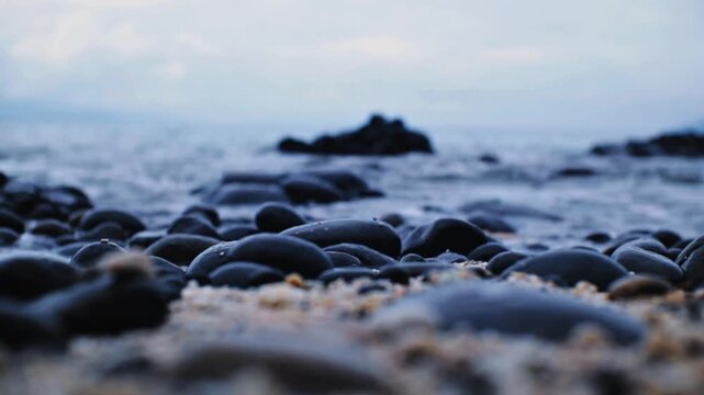 Close-up shot of smooth black basalt stones on a sandy shore as sea waves wash over them. Ideal for mood, mindfulness, coastal nature, volcanic beach, and calm background footage.