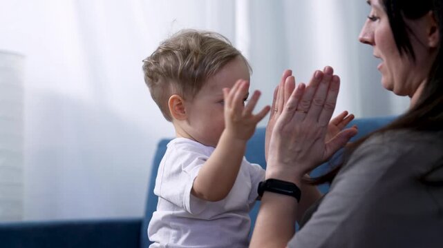 Mother and toddler play clapping hand game on sofa. Child claps palm with mom indoors. Toddler boy touches mother hand joyfully. Family bonding with child at home. Mom and baby playing together.