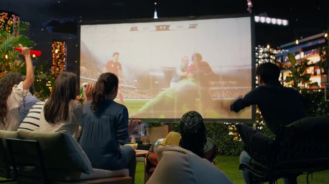 Zealous football supporters react joyfully to a goal during a projected game, sharing high fives as passionate spectators enjoy outdoor sports viewing.