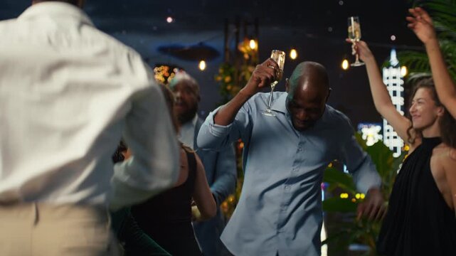Close up of joyful friends dancing at a nighttime rooftop party while holding champagne, with ecstatic partygoers moving to the rhythm of music and enjoying nightlife together above city lights.