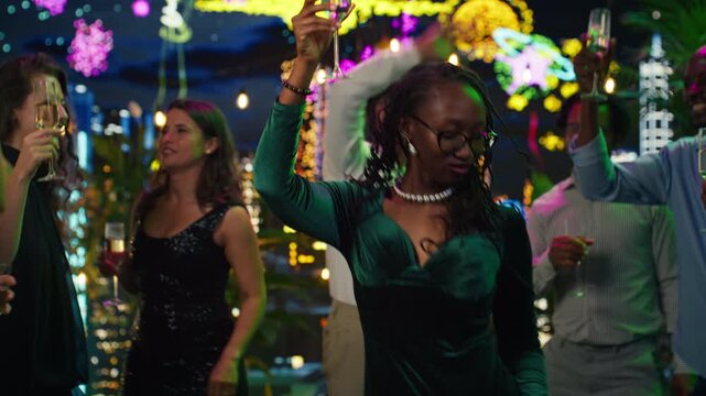 Smiling woman dances with friends on an illuminated rooftop terrace at night, drinking champagne during a festive New Year's Eve social event.