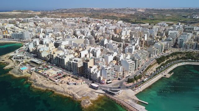 St Paul's Bay and Bugibba, Malta seafront with apartment blocks, marina, boats, promenade and turquoise Mediterranean Sea on a sunny spring day, captured by drone.