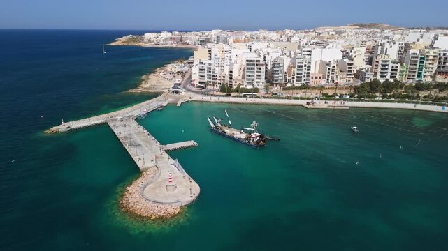 St Paul's Bay, Malta harbor with dredging vessels near breakwater construction, marina and waterfront apartment blocks along the Mediterranean coastline, filmed by drone.