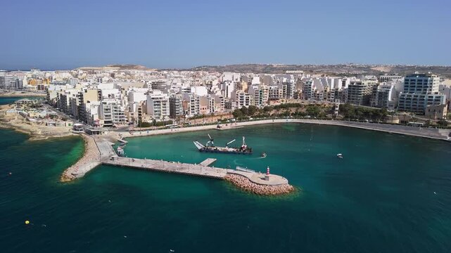 St Paul's Bay, Malta harbor with dredging vessel near Bugibba breakwater and jetty, clear Mediterranean water, urban waterfront and marina cityscape captured by drone.