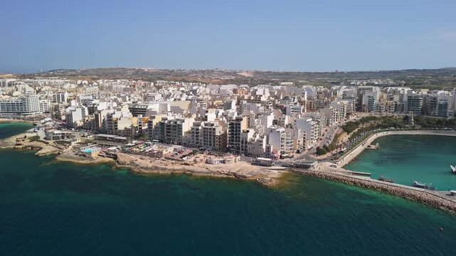 St Paul's Bay and Bugibba, Malta coastline with marina, promenade, apartment buildings, boats and turquoise Mediterranean water on a sunny spring day, captured by drone.