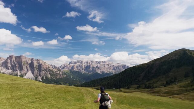 Dynamic tracking shot following hiker running over meadow crest toward towering rock walls in daylight, Dolomites, South Tyrol, Alps, Italy