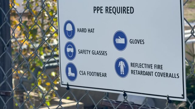 A PPE REQUIRED sign on a chain link fence listing hard hat, safety glasses, CSA footwear, gloves and reflective flame resistant coveralls.