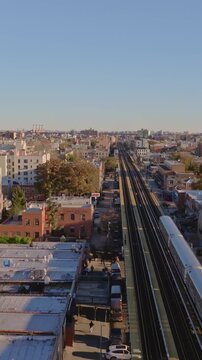 Vertical video: Elevated train traversing residential NYC neighborhood in Queens, revealing urban landscape with buildings and streets under bright sunlight from aerial perspective