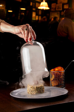 Smoking seafood tartare dish garnished with an octopus tentacle, with a blurred restaurant in the background.