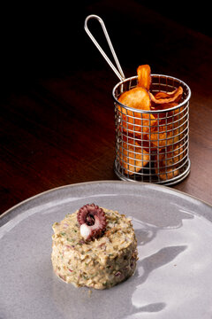 Close-up of seafood tartare garnished with a gourmet octopus tentacle, served with potatoes on a wooden table.